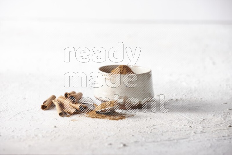 Ceramic beige bowl full of cinnamon powder and a metal spoon with cinnamon sticks next of it on a textured white background