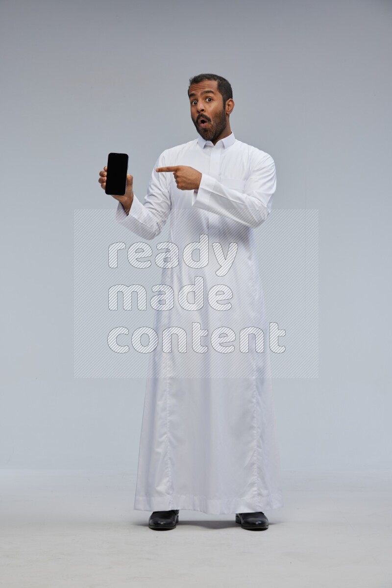 Saudi man wearing Thob standing showing phone to camera on Gray background