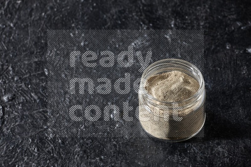 A glass jar full of white pepper powder on textured black flooring