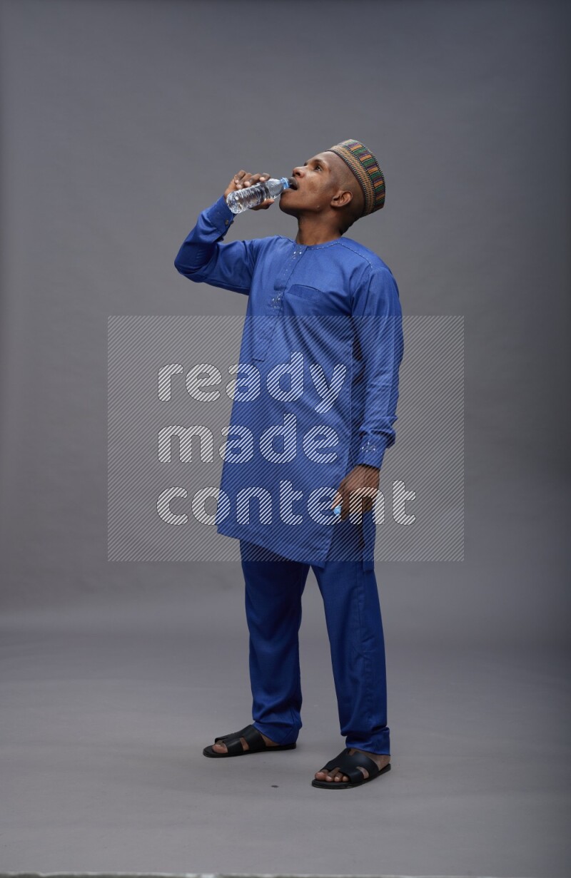 Man wearing Nigerian outfit standing drinking water on gray background
