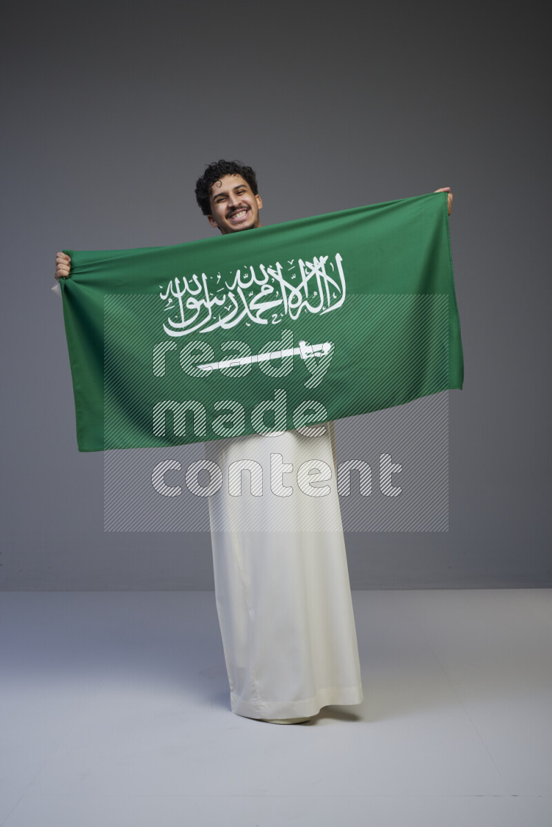 A Saudi man standing wearing thob holding big Saudi flag on gray background