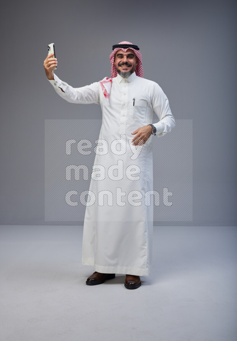 Saudi man Wearing Thob and red Shomag standing taking selfie on Gray background