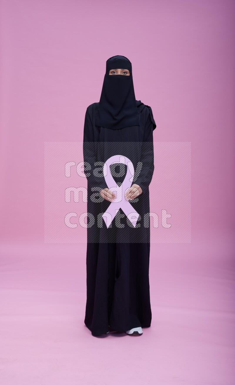 Saudi woman wearing abaya and niqab standing holding awareness ribbon on pink background