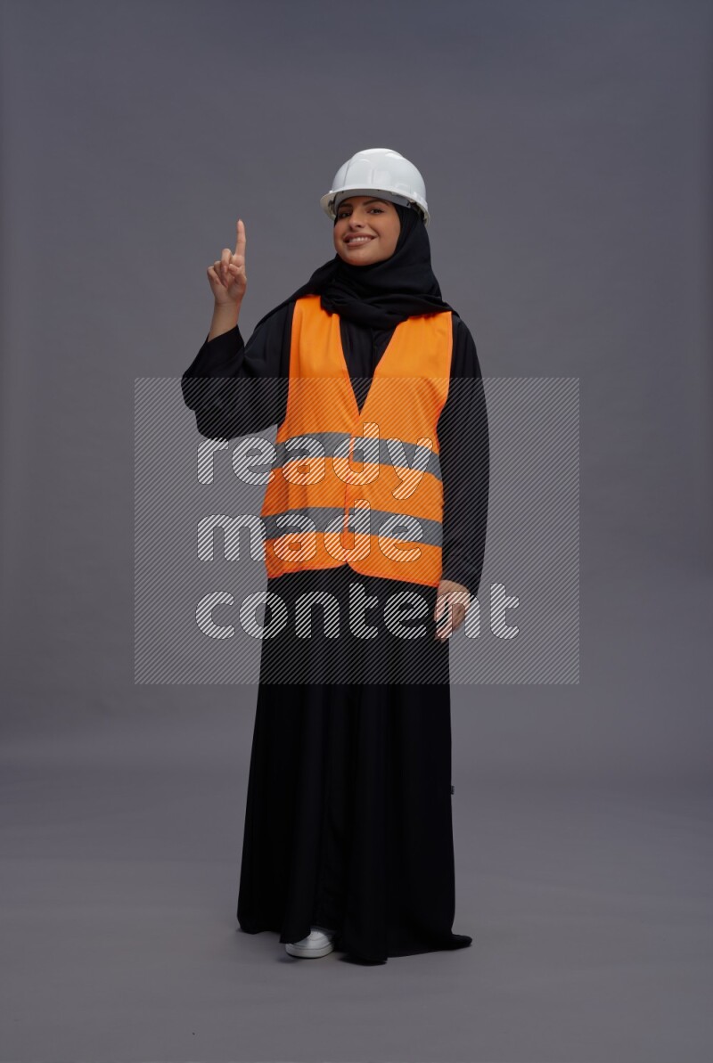 Saudi woman wearing Abaya with engineer vest standing interacting with the camera on gray background