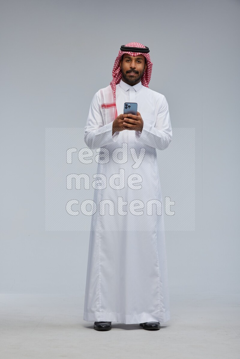 Saudi man Wearing Thob and shomag standing texting on phone on Gray background