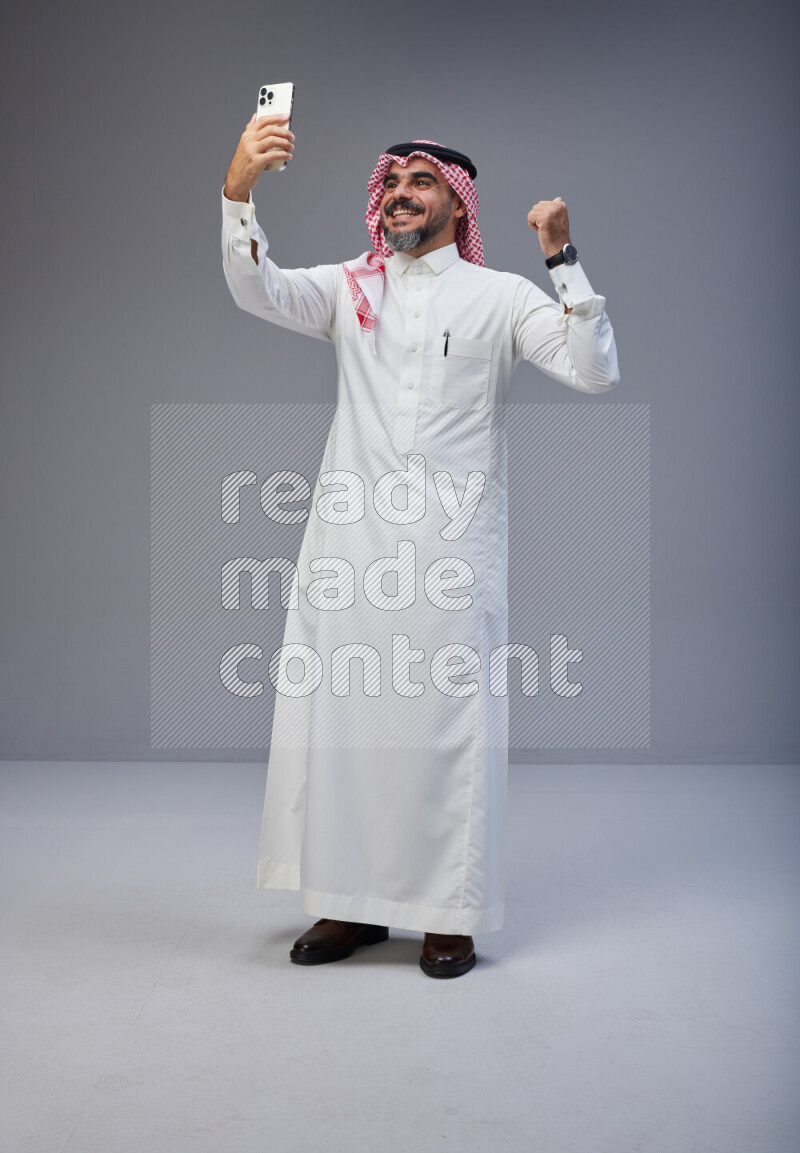 Saudi man Wearing Thob and red Shomag standing taking selfie on Gray background