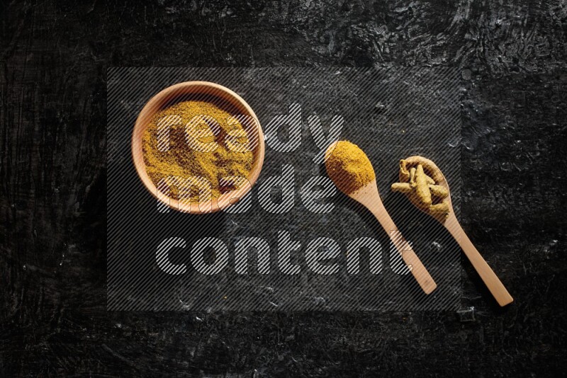 A wooden bowl full of turmeric powder and 2 wooden spoons full of dried turmeric finger and turmeric powder on textured black flooring