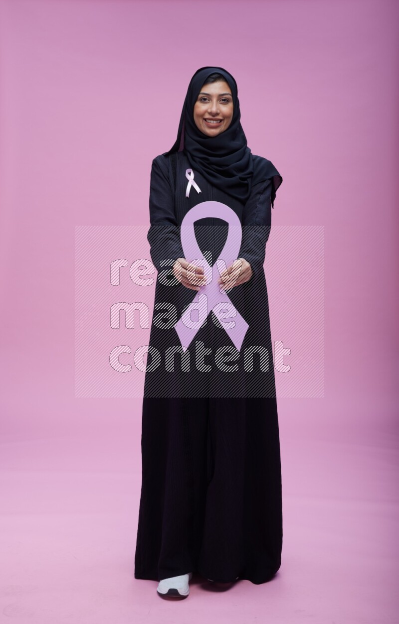 Saudi woman wearing pink ribbon on abaya standing holding awareness ribbon on plain pink background