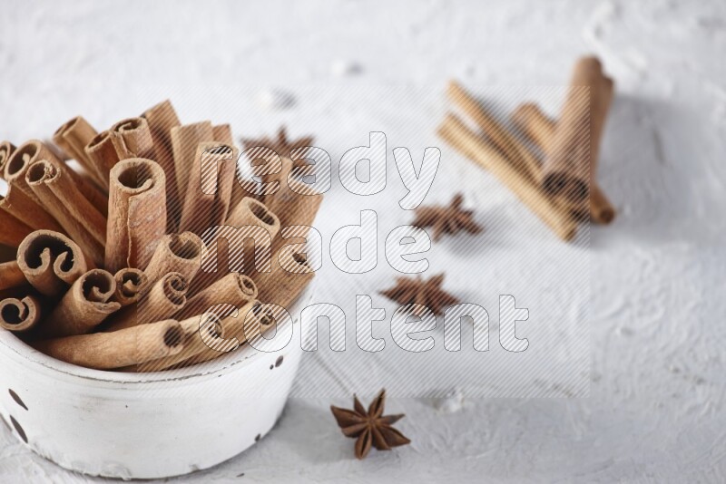 White bowl full of cinnamon sticks surrounded by star anis on a textured white background in different angles
