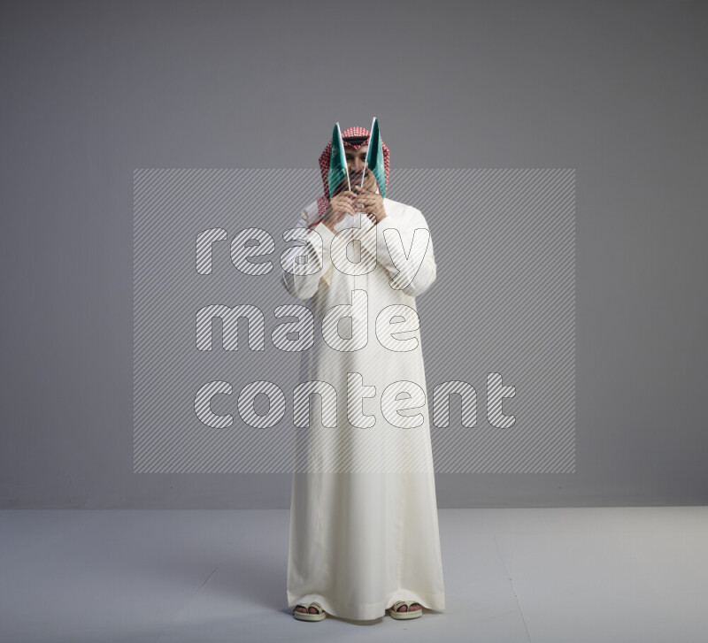 A Saudi man standing wearing thob and red shomag raising small Saudi flag on gray background