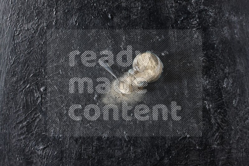 A flipped herbal glass jar and a metal spoon full of white pepper powder with spilled powder on textured black flooring