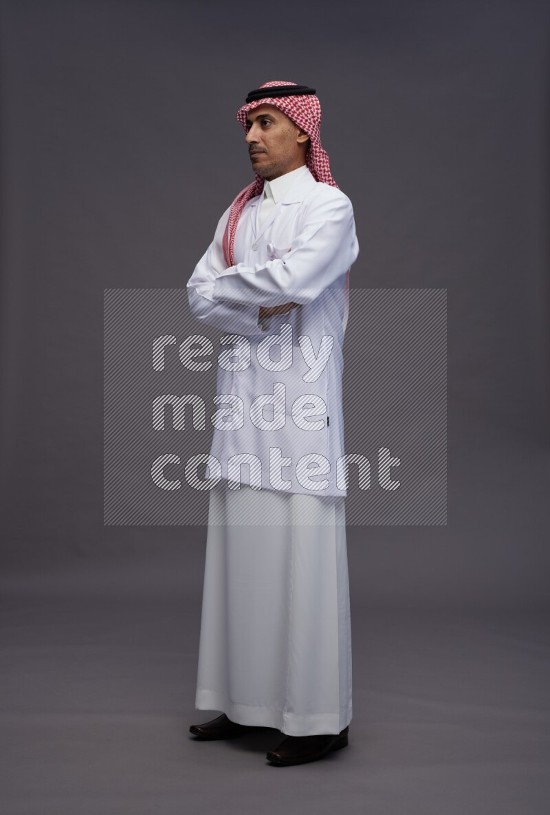 Saudi man wearing thob with lab coat and shomag with pocket employee badge standing with crossed arms on gray background