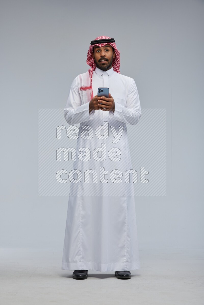 Saudi man Wearing Thob and shomag standing texting on phone on Gray background