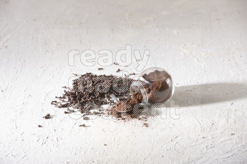 A flipped glass spice jar and a metal spoon full of cloves powder and powder came out of the jar with cloves spread on textured white flooring