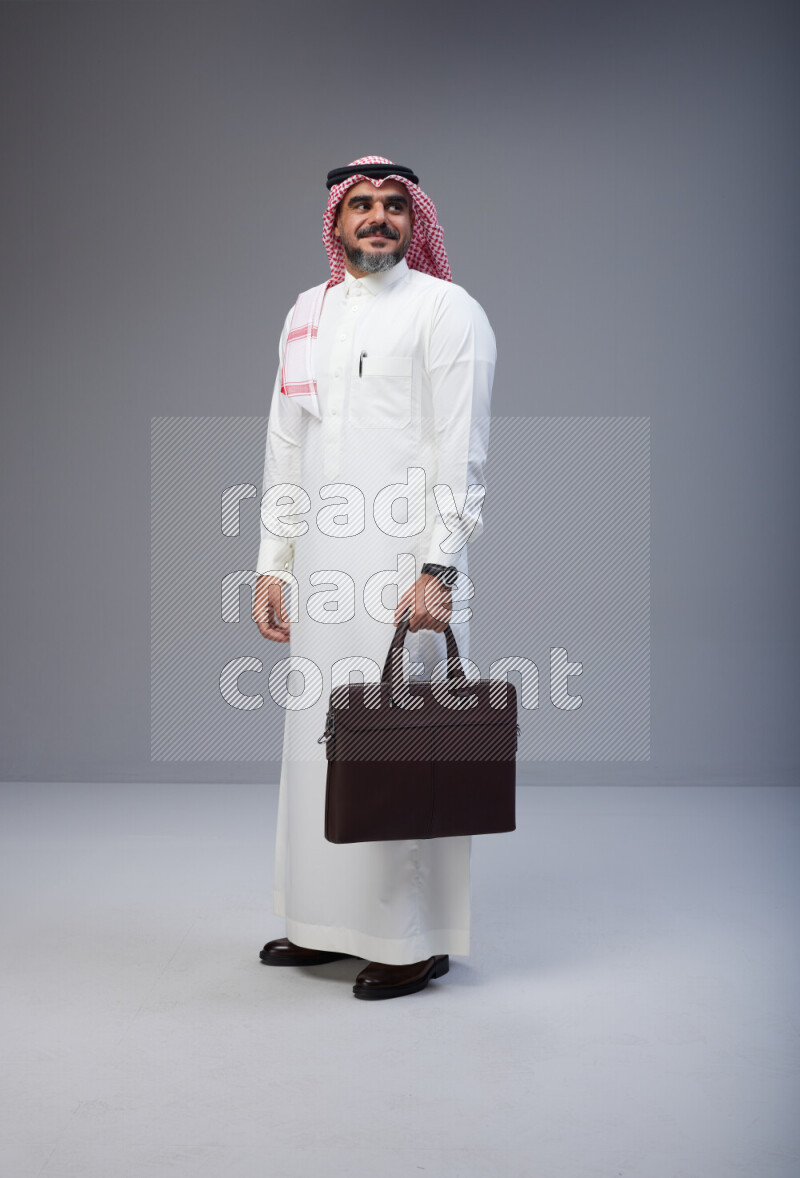 Saudi man Wearing Thob and red Shomag standing holding bag on Gray background