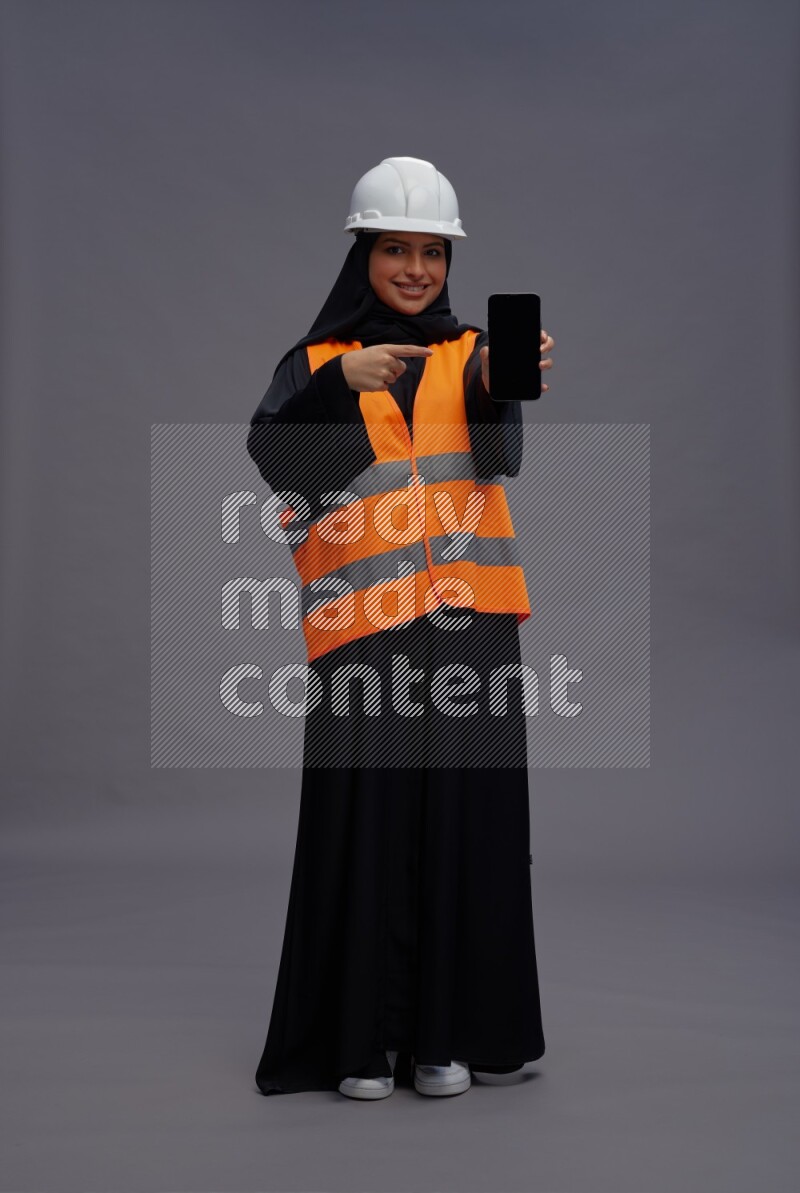 Saudi woman wearing Abaya with engineer vest standing showing phone to camera on gray background