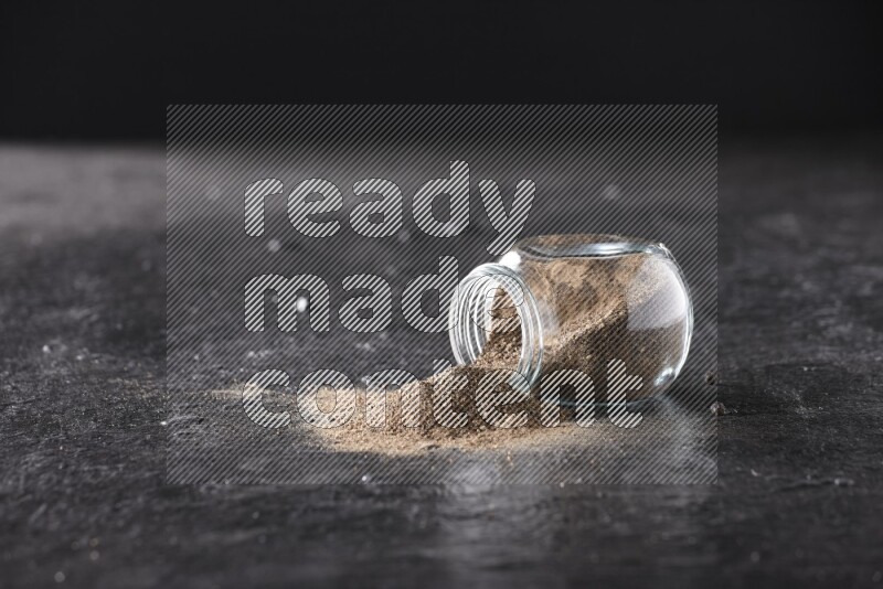A flipped glass spice jar full of black pepper powder on textured black flooring