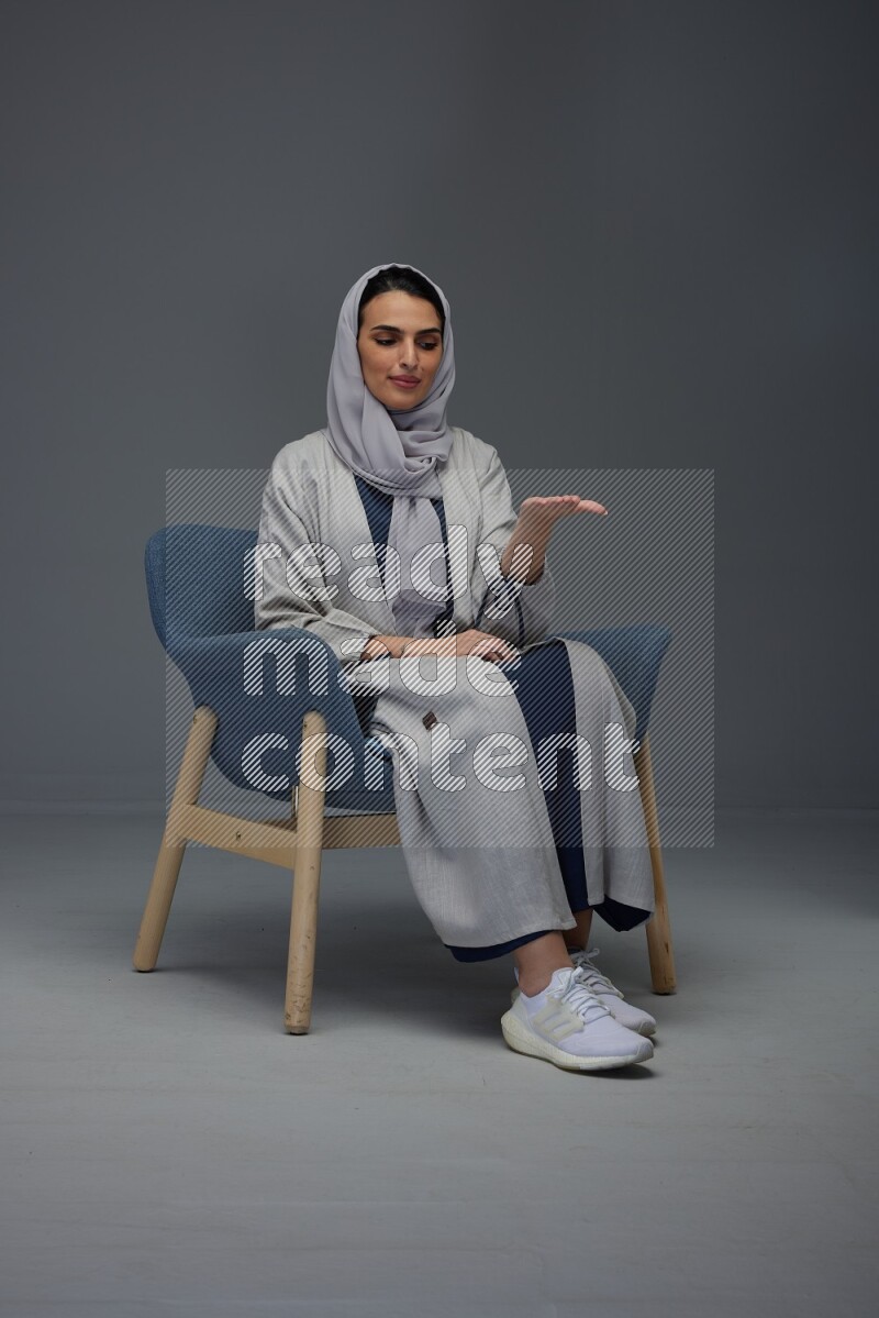 A Saudi woman wearing a light gray Abaya and head scarf sitting on a dark grey chair while making multi hand gestures eye level on a grey background