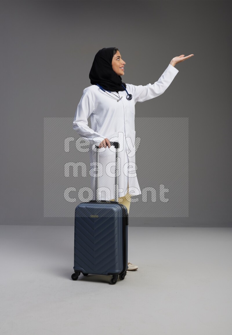 Saudi woman wearing lab coat with stethoscope standing holding Travel bag on Gray background