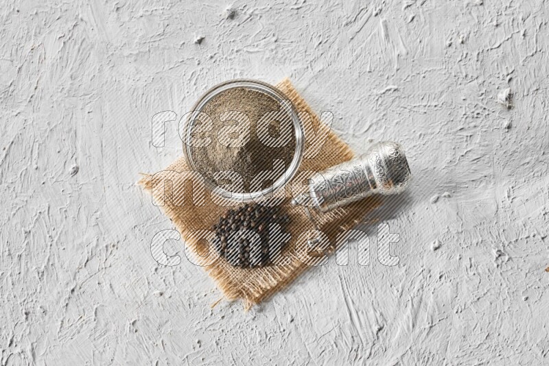 A glass bowl full of black pepper powder, black pepper beads and a turkish metal grinder on burlap fabric on textured white flooring