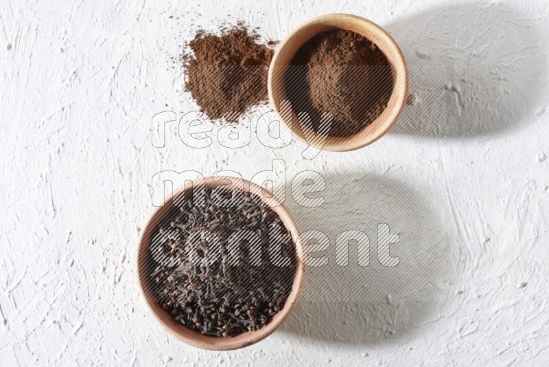 2 wooden bowls full of cloves powder and whole cloves on a textured white flooring