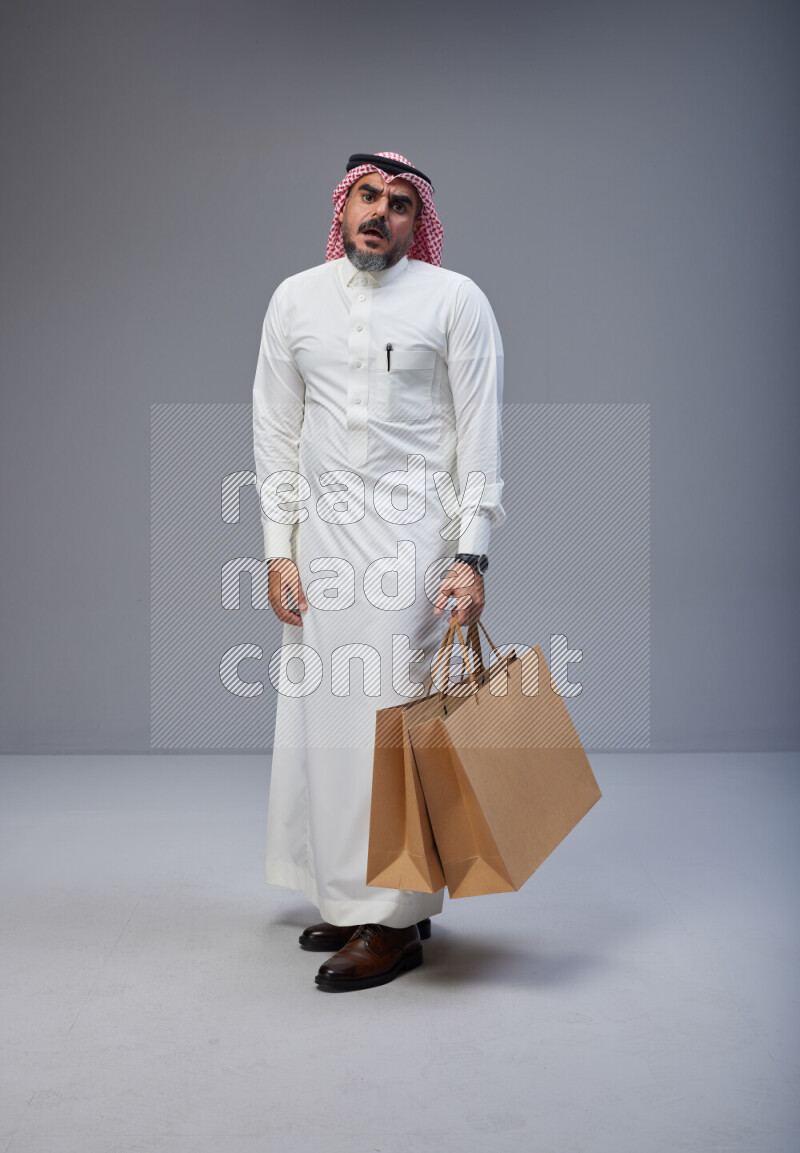 Saudi man Wearing Thob and red Shomag standing holding shopping bag on Gray background