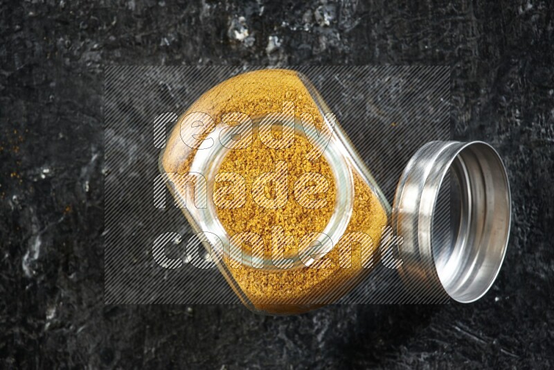 A glass spice jar full of turmeric powder on a textured black flooring
