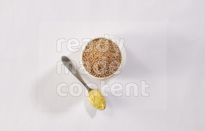 A beige pottery bowl full of mustard seeds and a metal spoon full of mustard paste on white flooring