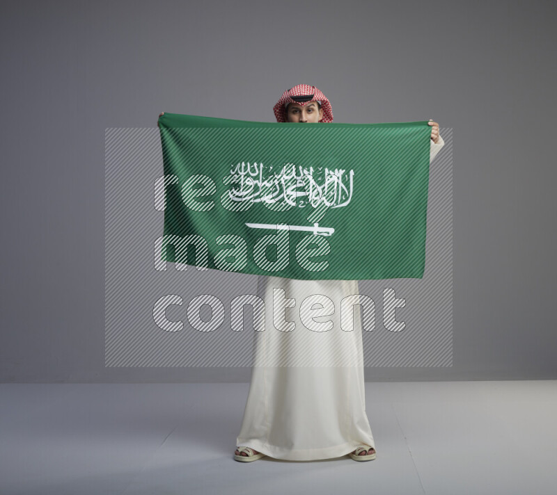 A saudi man standing wearing thob and red shomag holding big saudi flag on gray background