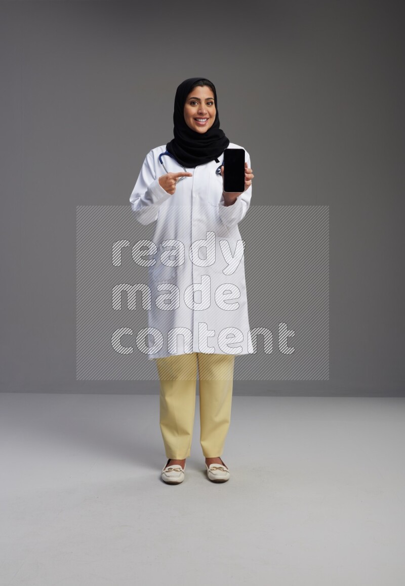 Saudi woman wearing lab coat with stethoscope standing showing phone to camera with sign in the back on Gray background