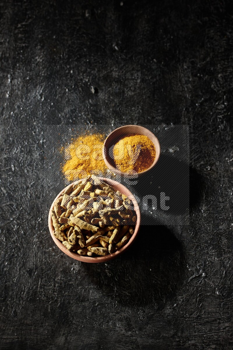 2 wooden bowls, one full of turmeric powder and the other full of dried turmeric whole fingers on a textured black flooring