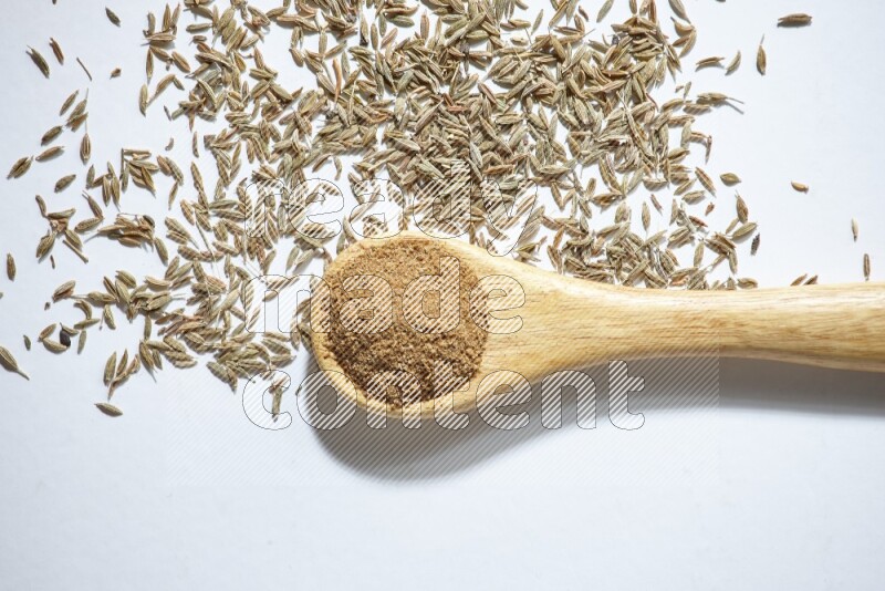 A wooden spoon full of cumin powder and cumin seeds beside it on white flooring