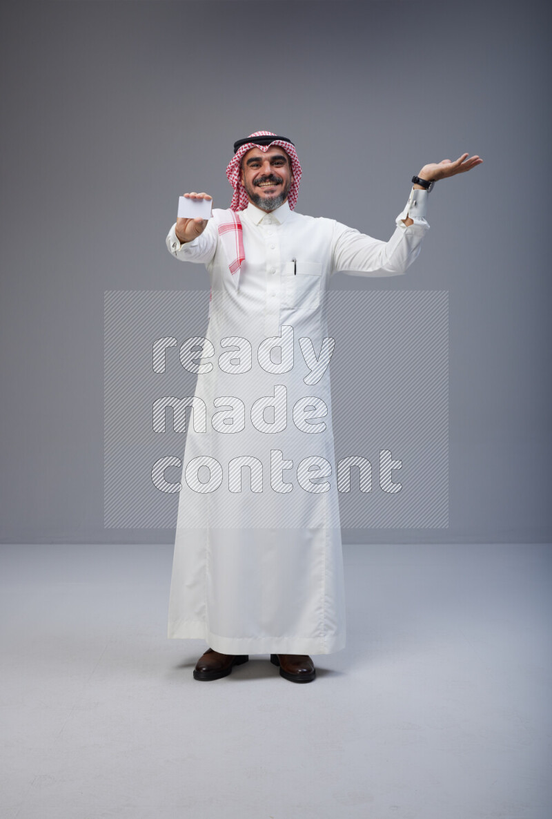 Saudi man Wearing Thob and red Shomag standing holding ATM card on Gray background