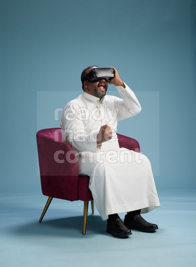 Saudi Man without shimag sitting on chair  gaming with VR on blue background