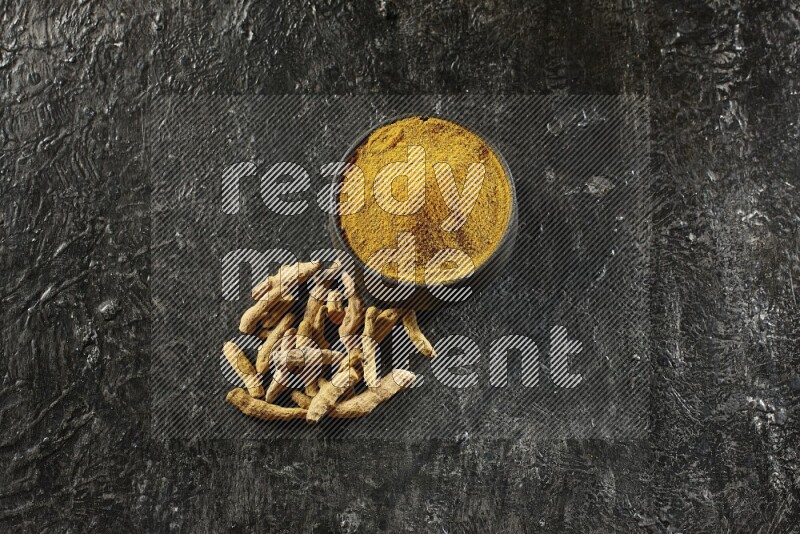 A black pottery bowl full of turmeric powder and dried turmeric whole fingers next of it on textured black flooring