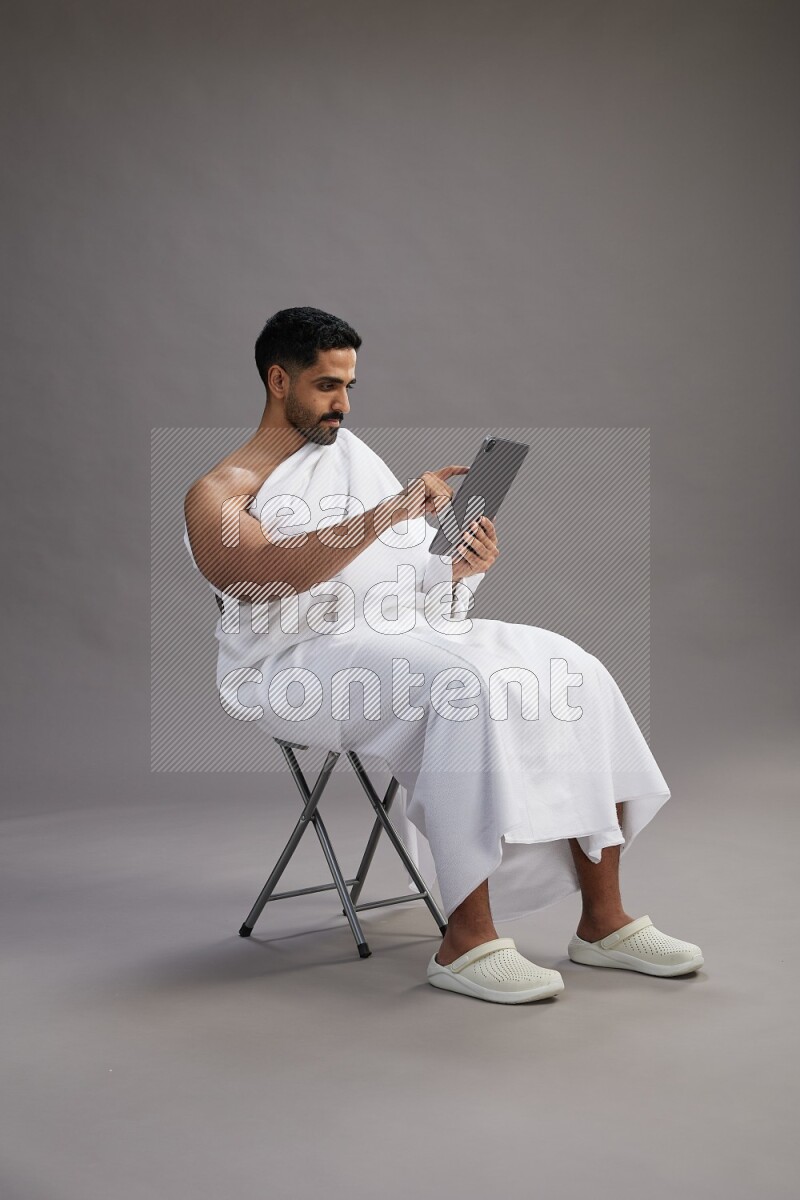 A man wearing Ehram sitting on chair holding a tablet on gray background