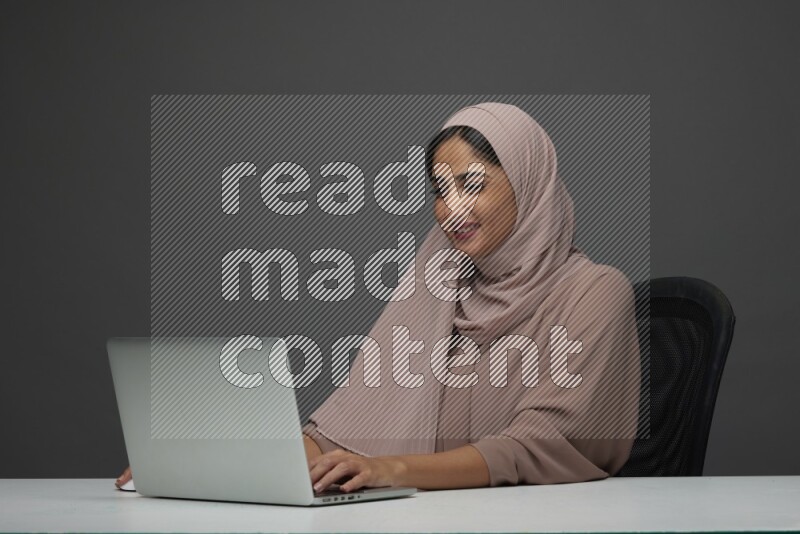 A Saudi woman Setting on her desk Typing on her laptop on a Gray Background wearing Brown Abaya with Hijab