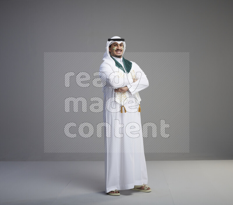 A Saudi man standing wearing thob and white shomag with face painting and Saudi flag scarf on gray background