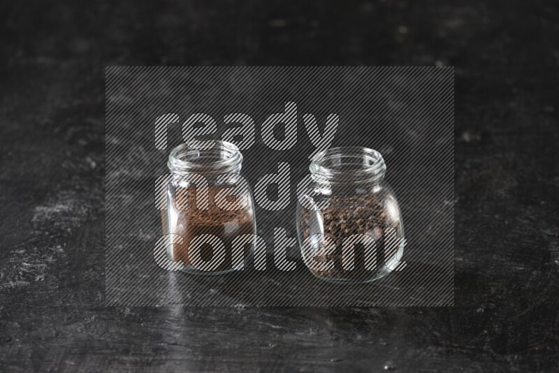 Glass spice jars full of cloves and cloves powder on textured black flooring