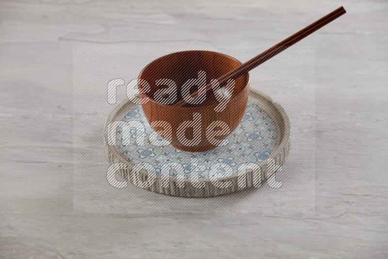 brown wood round bowl on top of multi color round ceramic plate and chopsticks, on grey textured countertop