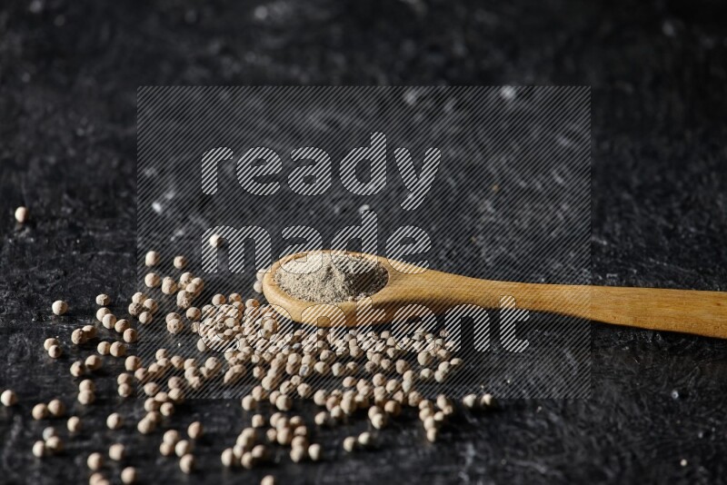 A wooden spoon full of white pepper powder with white pepper beads on textured black flooring