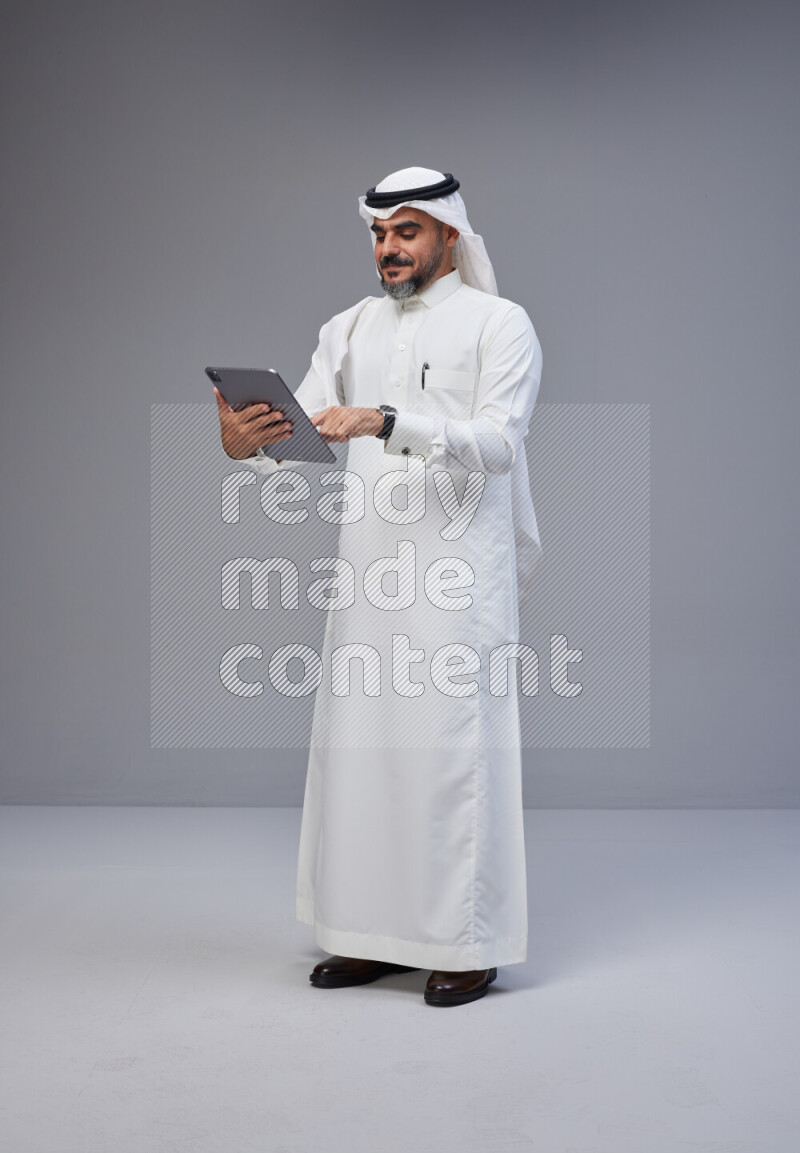 Saudi man Wearing Thob and white Shomag standing working on tablet on Gray background