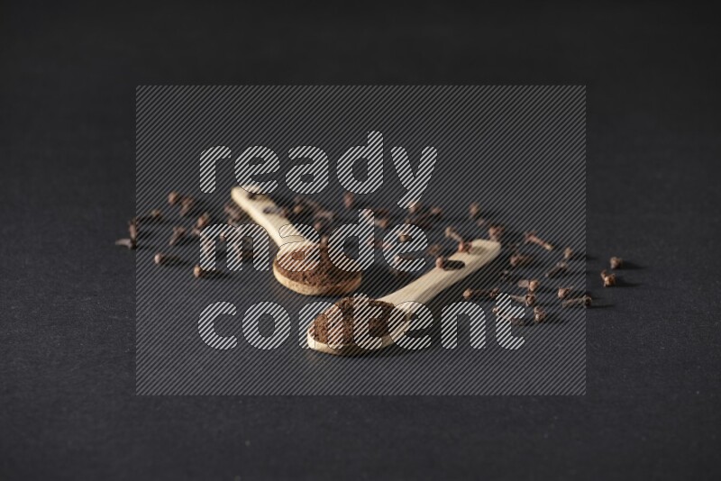 ملعقتان خشبيتان ممتلئتان ببودرة القرنفل مع حبوب القرنفل المتناثرة على خلفية سوداء