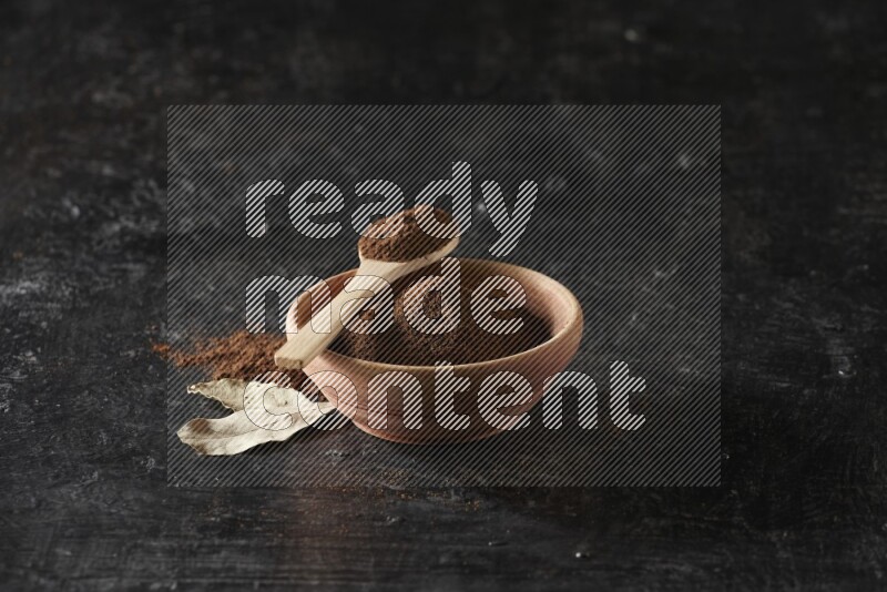 A wooden bowl and a wooden spoon full of cloves powder with laurel leaves on a textured black flooring