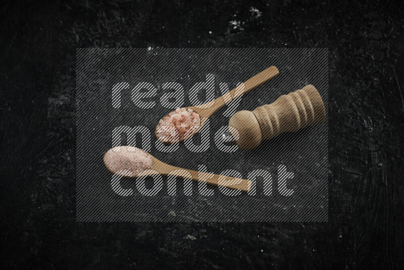 2 wooden spoons filled with fine and coarse salt with wooden grinder beside them on black background