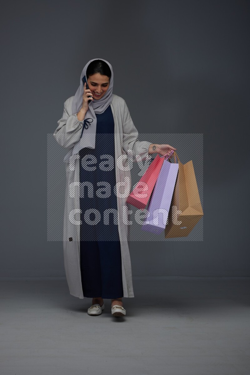 A Saudi woman wearing a light gray Abaya and head scarf standing and holding shopping bags making different poses eye level on a grey background