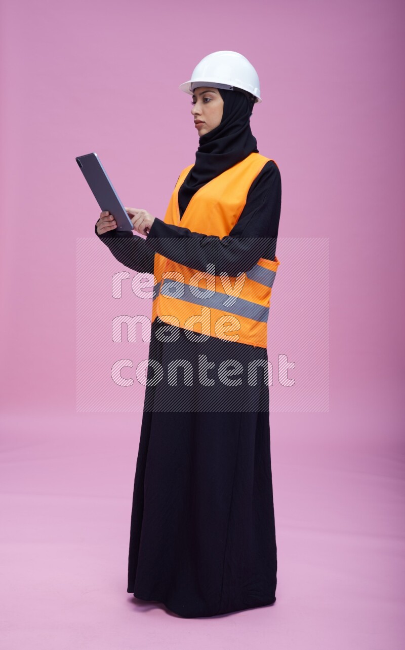 Saudi woman wearing Abaya with engineer vest and helmet standing working on tablet on pink background