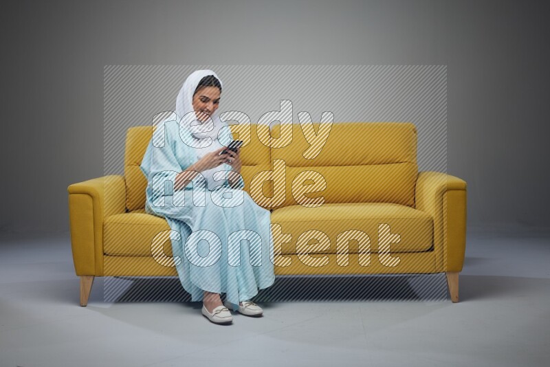 A Saudi woman wearing a light blue Abaya and white head scarf sitting on a yellow sofa and using her phone eye level on a grey background