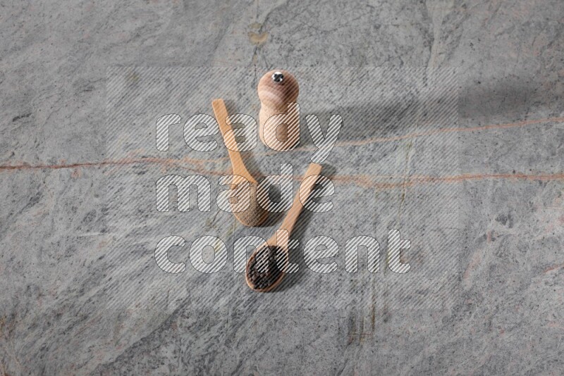 ملعقتان خشبيتان، ملعقة ممتلئة بمسحوق الفلفل الأسود والأخرى ممتلئة بحبات الفلفل الأسود ومطحنة خشبية على خلفية رخام رمادية