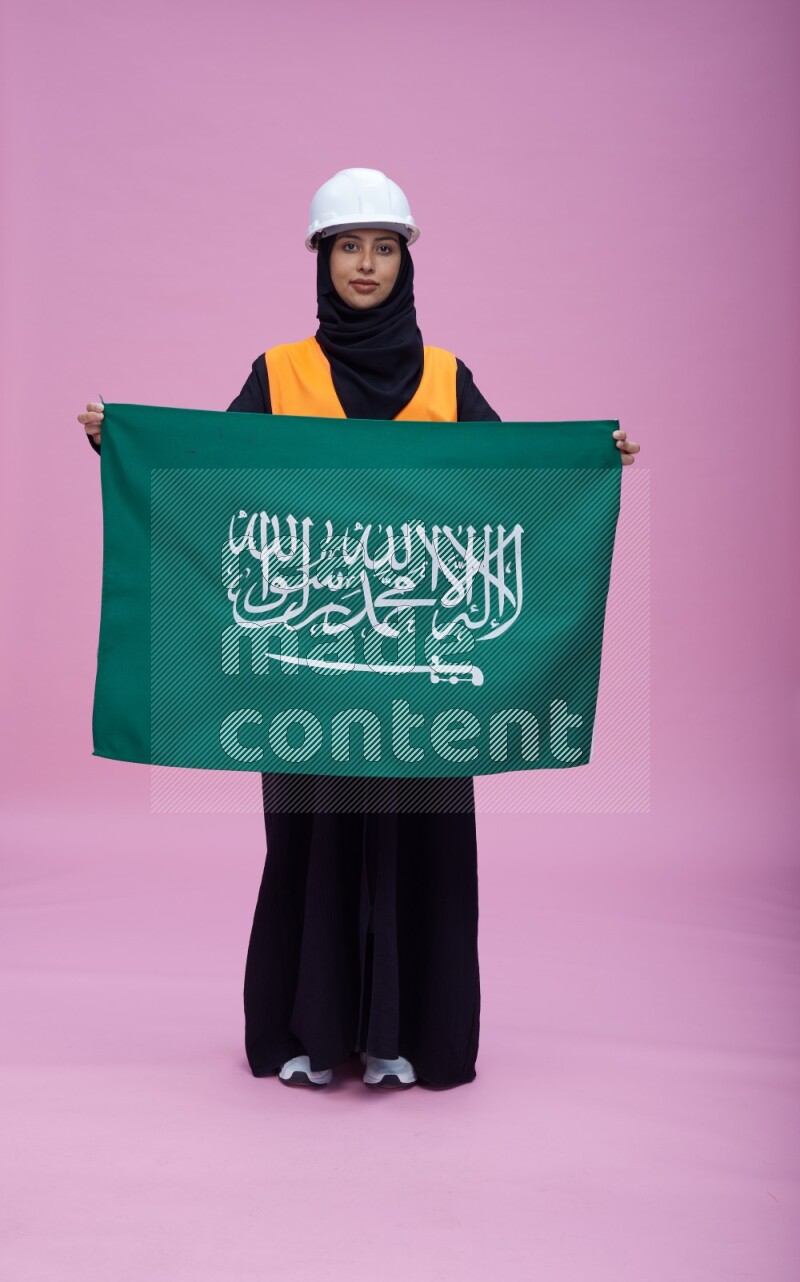 Saudi woman wearing Abaya with engineer vest and helmet standing holding Saudi flag on pink background
